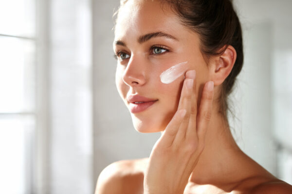 A calm young woman gently applying rich face cream to her healthy glowing skin in a bright modern bathroom.